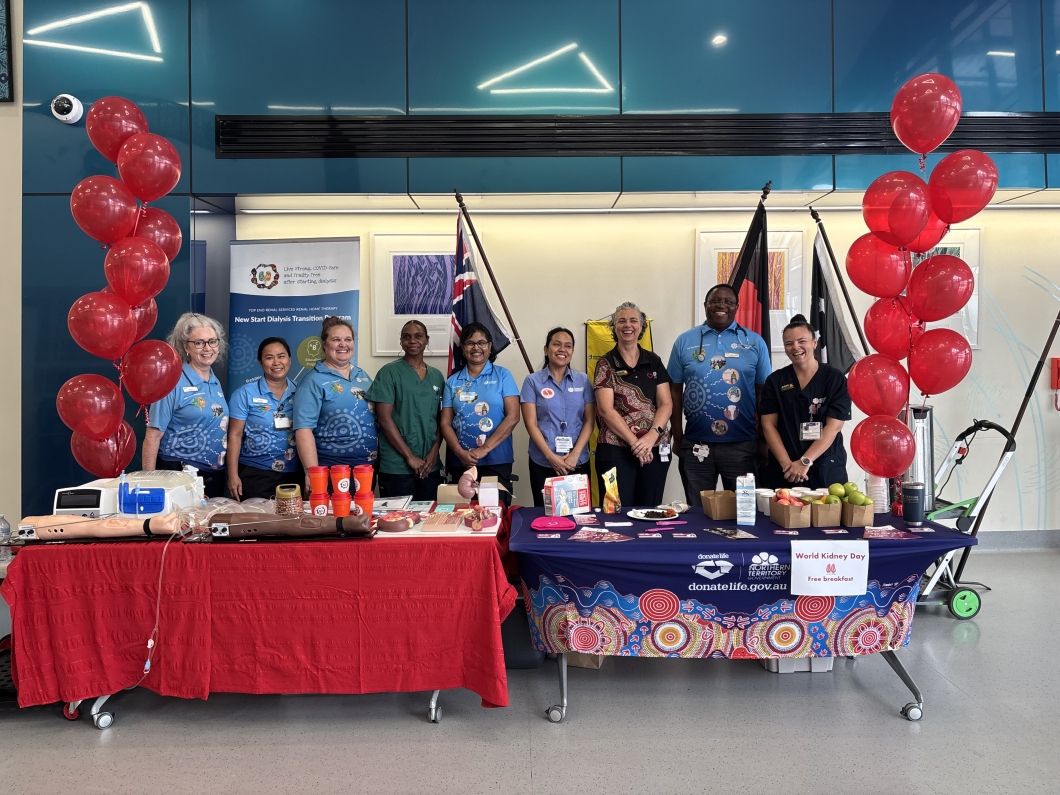 A group of 9 healthcare staff stand behind 2 display tables at an indoor World Kidney Day event. The tables are covered with educational materials, models of kidneys and dialysis equipment, pamphlets, cups, and fruit. Red balloons frame both sides of the display. Behind the group are banners and several flags, including Aboriginal and Torres Strait Islander flags, along with a sign reading ‘World Kidney Day – Free breakfast’ and ‘donatelife.gov.au’.