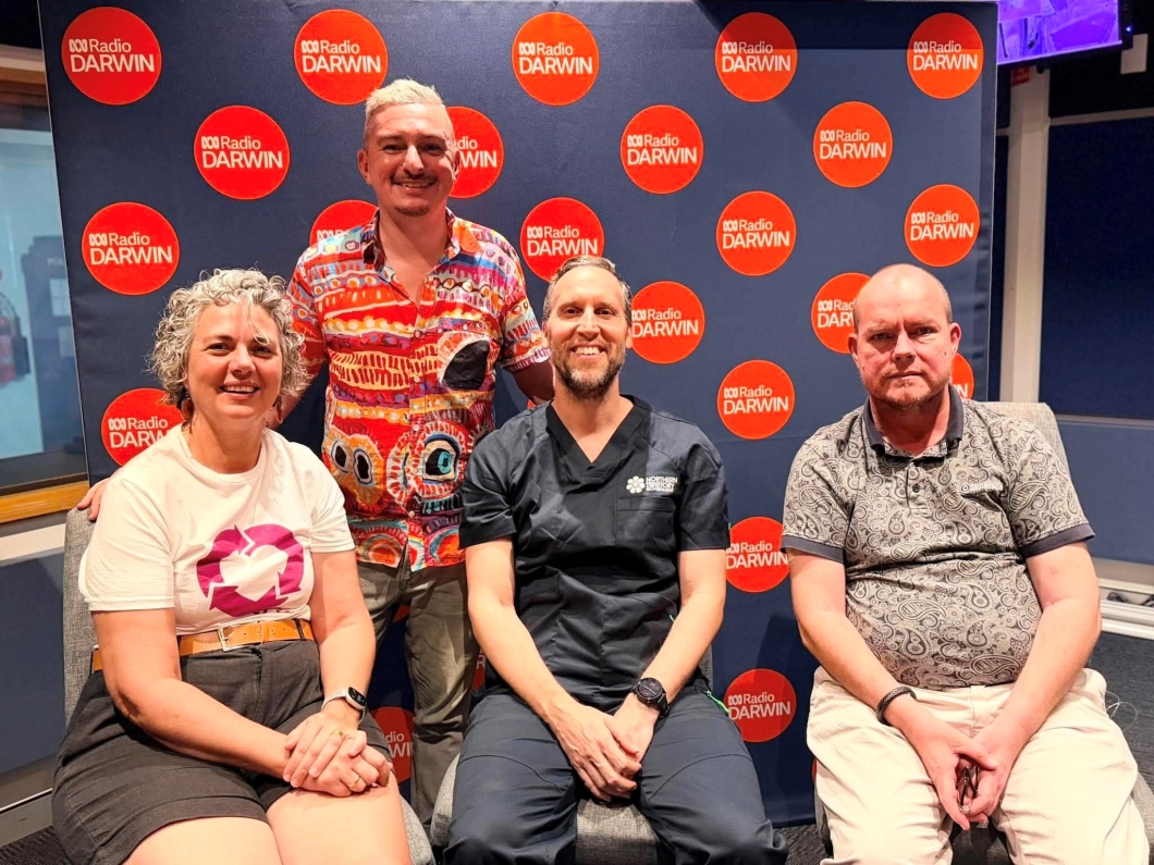 4 people pose for a group photo inside an ABC Radio Darwin studio. One person wears a DonateLife shirt featuring the magenta DonateLife logo, while another wears clinical scrubs. Behind the group is a backdrop displaying multiple ABC Radio Darwin logos, with studio equipment and broadcasting facilities visible around the room.