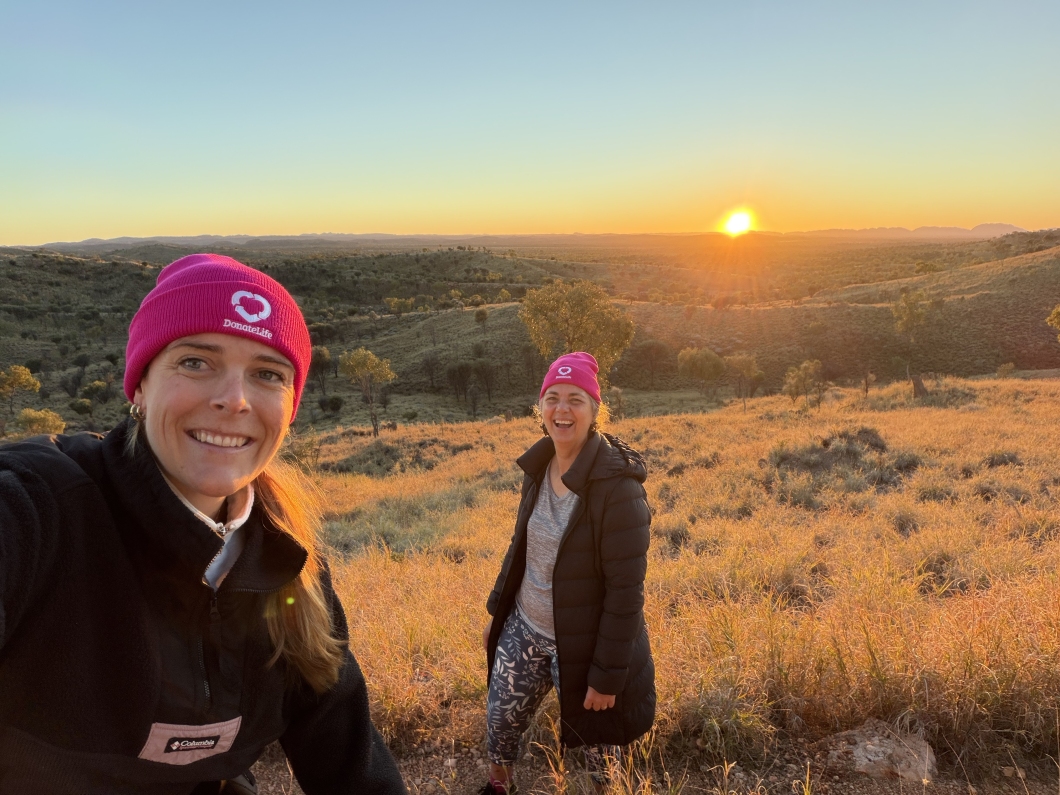 2 people wearing magenta DonateLife beanies stand on a hillside at sunrise, with rolling outback landscape stretching to the horizon behind them. Golden morning light illuminates the dry grass, scattered trees and distant ranges. One person takes a selfie while the other stands further back, both wearing DonateLife-branded beanies against the backdrop of a clear sky and rising sun.