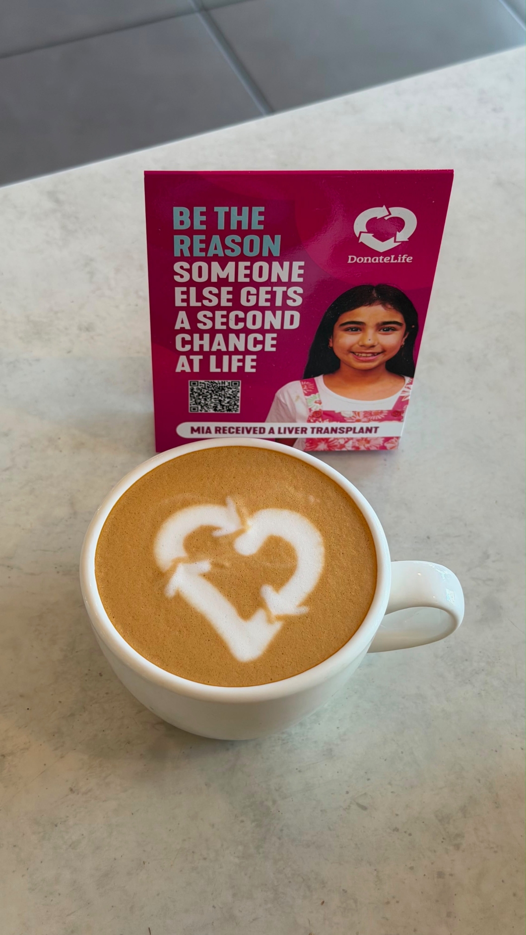 A cup of coffee sits on a café table with the DonateLife logo created in the milk foam. Behind the coffee is a pink DonateLife promotional card featuring a transplant recipient and the message, ‘Be the reason someone else gets a second chance at life’. A QR code and DonateLife branding are also visible on the card.