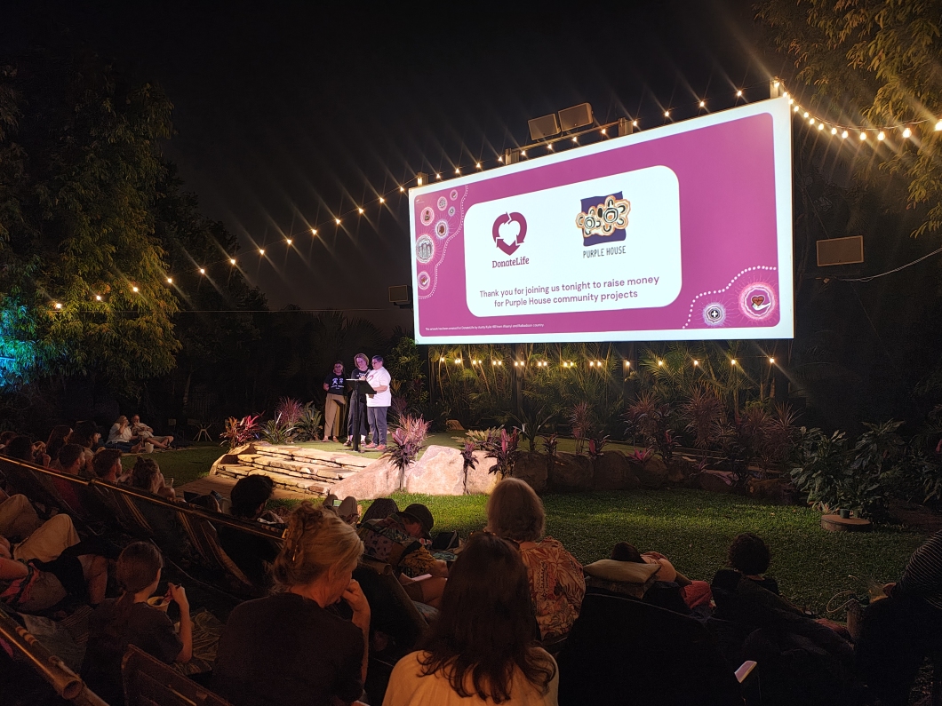 An outdoor nighttime event with a large screen displaying logos for DonateLife and Purple House. The screen reads, 'Thank you for joining us tonight to raise money for Purple House community projects.' Attendees are seated on grass and chairs, watching 2 speakers on a small stage. String lights overhead add to the festive ambiance. 