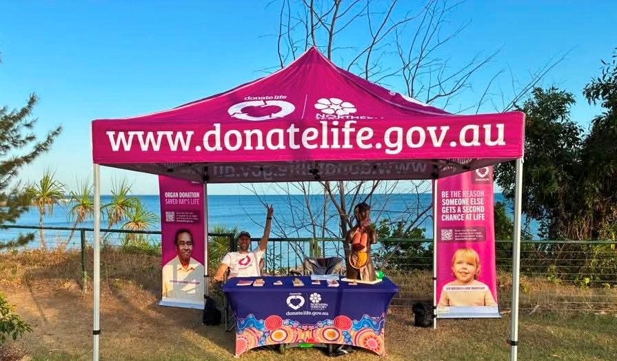 A DonateLife information stall is set up under a magenta marquee on the Nightcliff Foreshore, overlooking the ocean. The marquee displays the DonateLife website address and is flanked by banners promoting organ donation. A table covered with DonateLife information and promotional materials sits beneath the shelter, with volunteers standing behind it. Coastal vegetation, a fence and the sea are visible in the background under a clear blue sky.