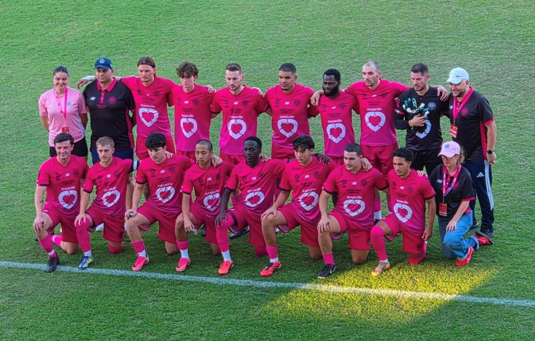 A football team and coaching staff pose for a group photo on the field before a match, wearing DonateLife-branded magenta jerseys featuring the DonateLife heart logo. The players stand and kneel in two rows on the grass, with coaches and event supporters positioned at either end of the group. Several people wear matching magenta lanyards and caps, highlighting support for organ and tissue donation awareness.