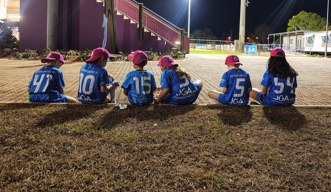 6 children wearing blue ‘AZZURRI UNITED FC’ jerseys and magenta caps sit on the ground with their backs to the camera at an outdoor sports facility at night. 