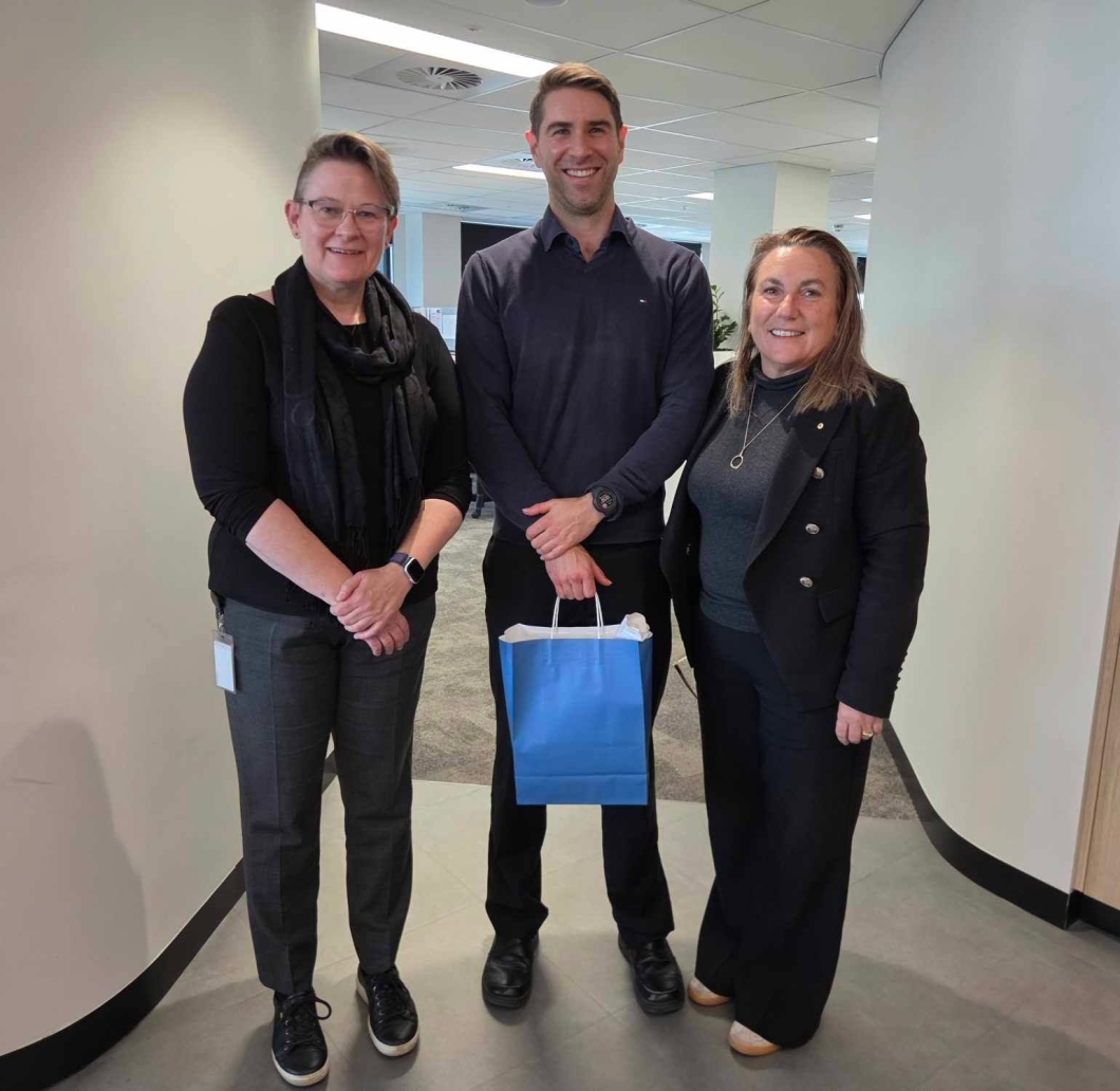 3 people stand together in an office corridor for a farewell photo. The person in the centre holds a blue gift bag, while the group poses in a modern workplace setting with desks and meeting areas visible in the background.