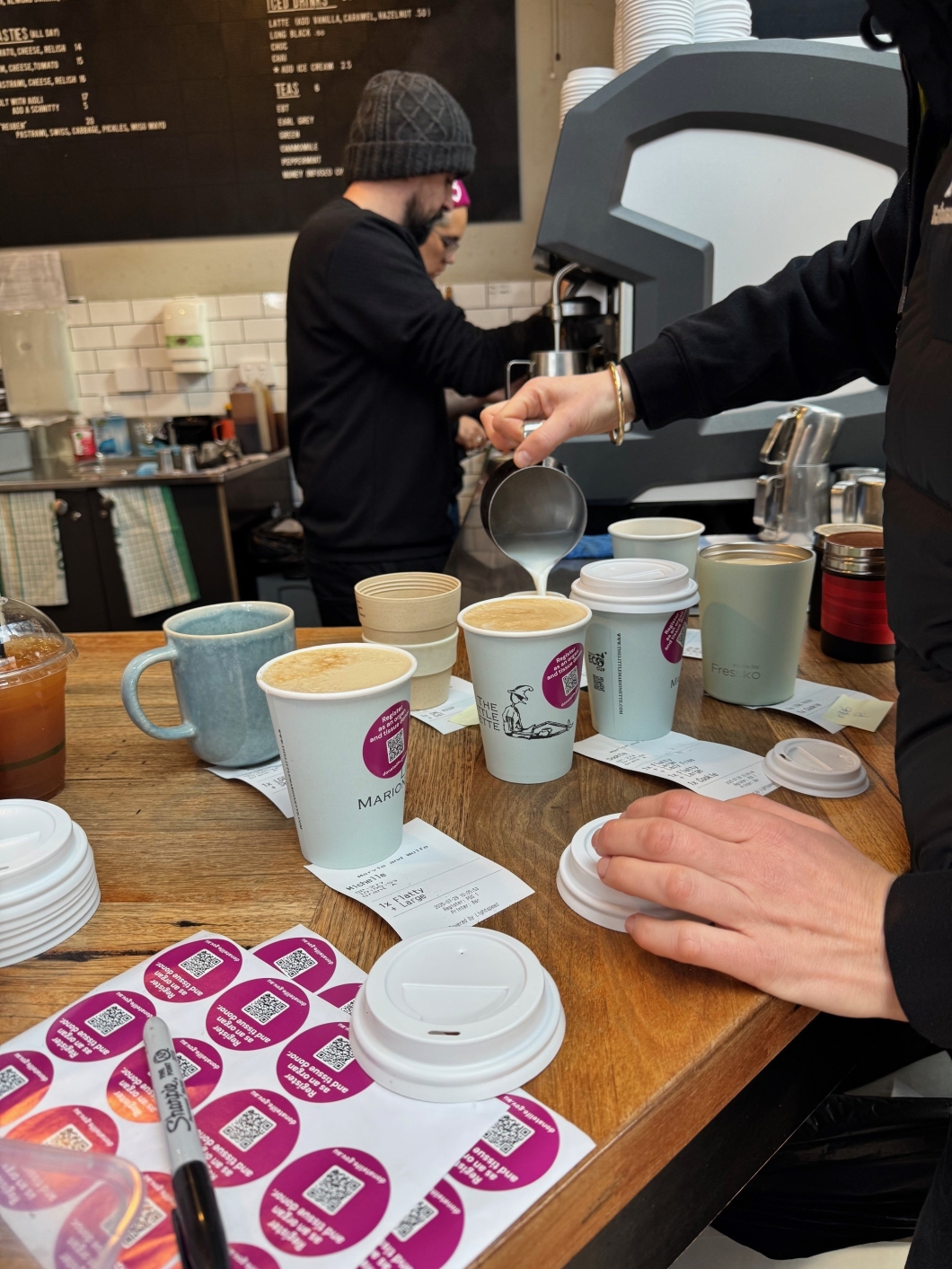 Two baristas working behind the counter of a café, one pouring milk into a coffee cup while another prepares drinks. The counter is filled with different coffee cups, lids, receipts, and stickers.