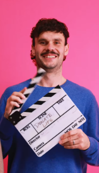 A person wearing a blue sweater holding a clapperboard with ‘DONATELIFE’ written on it, standing in front of a pink background.