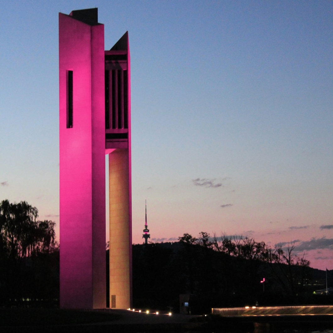 A tall, modern tower illuminated in pink light at dusk, featuring vertical slits and an open section near the top. A telecommunications tower and trees are silhouetted in the background against a twilight sky.