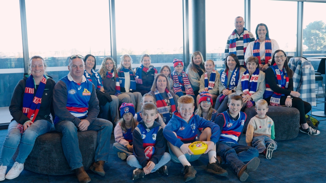 A group of adults and children wearing Western Bulldogs AFL team merchandise, including scarves, jerseys, and hats, gathered in a room with large windows and an outdoor view. Some children are holding footballs.