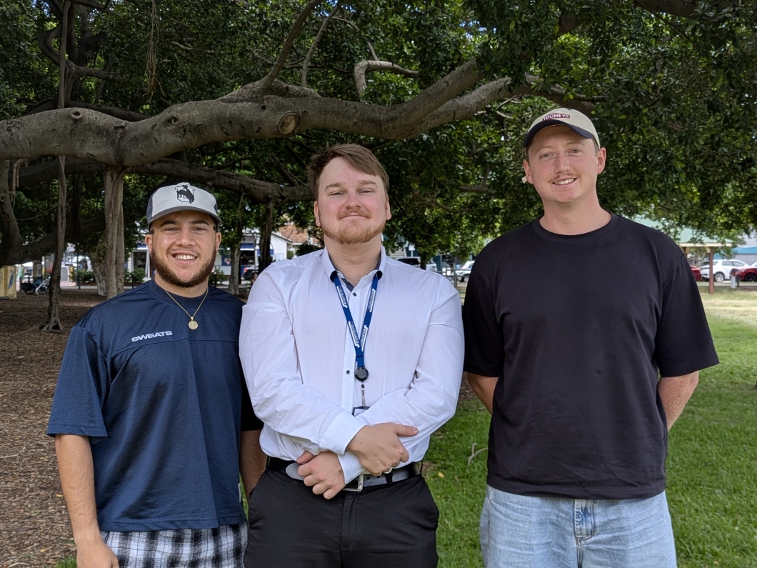 3 people stand together on a grassy area beneath large, spreading tree branches. One person is wearing a dark shirt and cap, another is dressed in a white collared shirt with a lanyard, and the third is wearing a black T‑shirt and light jeans. Parked cars and more trees are visible in the background.