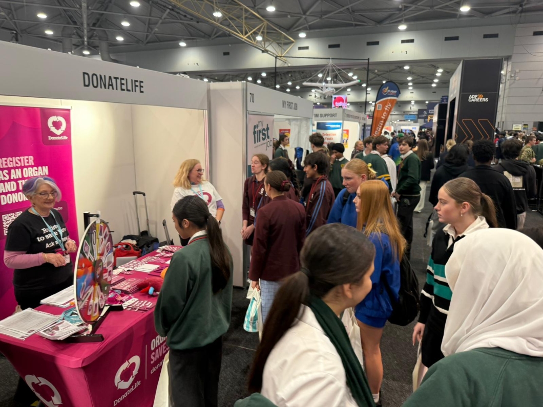 A busy indoor exhibition or career fair with multiple booths and attendees. The foreground features a bright pink 'DONATELIFE' booth with a banner reading 'REGISTER AS AN ORGAN AND TISSUE DONOR' and a QR code. A person behind the table wears a shirt that says 'REGISTER TO SAVE LIVES. ASK ME HOW.' Pamphlets and brochures are displayed on the table, and people are engaging with the booth. Other booths are visible in the background. 