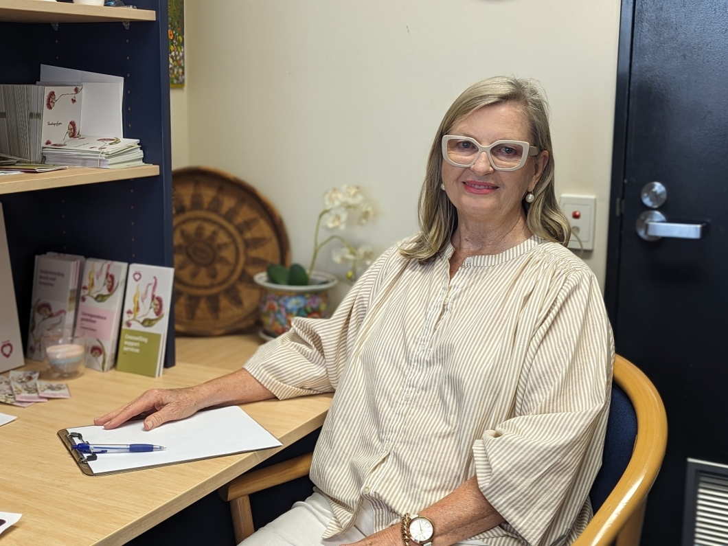 A person sits in an office-style room at a small desk. They are wearing a light, loose-fitting striped top and holding a clipboard with papers on the desk. Shelves beside them display brochures and books, and a small plant and decorative wooden plate sit on a side table in the background. A closed door is visible behind them.