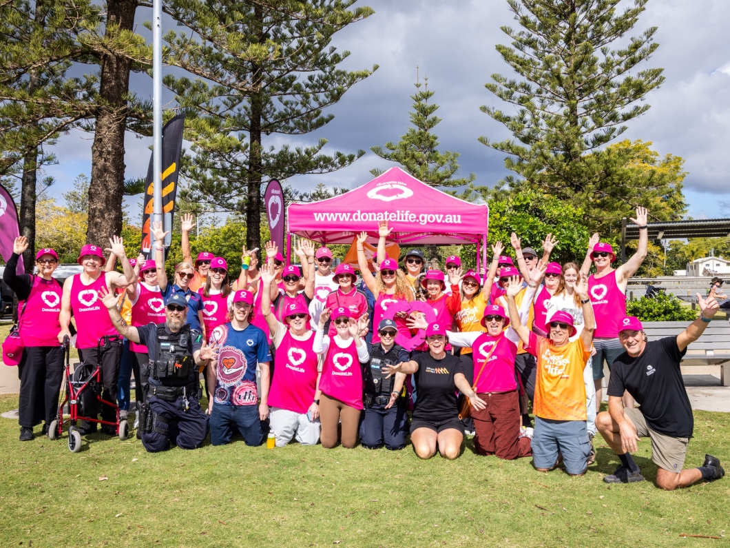 A large group of DonateLife volunteers, community members and local emergency services personnel gather in front of a magenta DonateLife marquee, raising their hands during an outdoor organ and tissue donation awareness event.