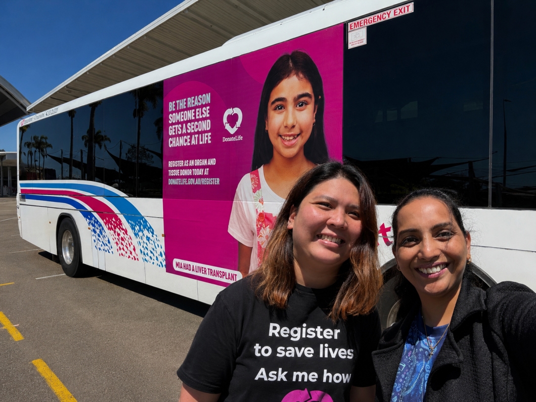 2 community outreach representatives stand beside a bus featuring a large magenta DonateLife advertisement encouraging people to register as organ and tissue donors.