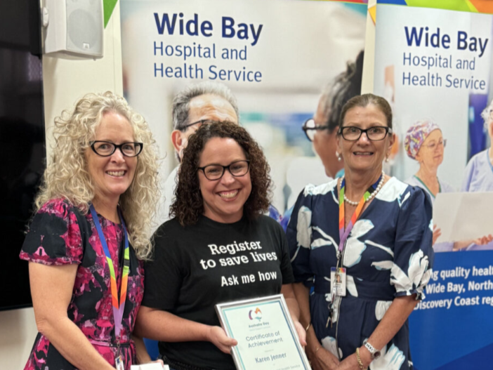 3 people stand together in front of two Wide Bay Hospital and Health Service banners. One person in the centre holds a framed Certificate of Achievement. The group is standing indoors, and the banners behind them display the Wide Bay Hospital and Health Service logo and images of healthcare settings.