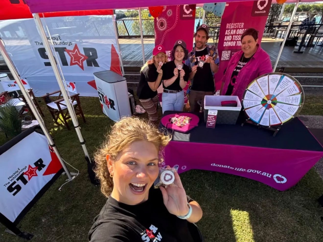 A community member takes a selfie at a DonateLife information stall, while volunteers stand behind a table decorated in magenta branding and share organ and tissue donation information at a local event.