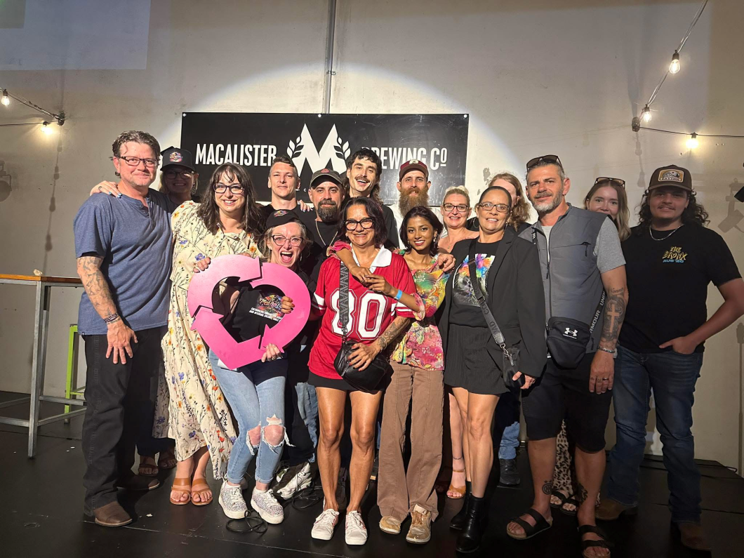 A group of people stand together on a stage in front of a sign reading ‘Macalister Brewing Co’, posing for a group photo at an indoor event. One person at the front holds a large magenta DonateLife heart-shaped logo prop, while others stand closely behind under warm string lights.