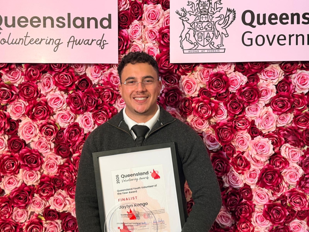 A person stands in front of a pink floral backdrop featuring ‘Queensland Volunteering Awards’ and ‘Queensland Government’ signs, holding a framed certificate. The certificate reads ‘2026 Queensland Volunteering Awards’ and ‘Queensland Youth Volunteer of the Year Award – Finalist’.
