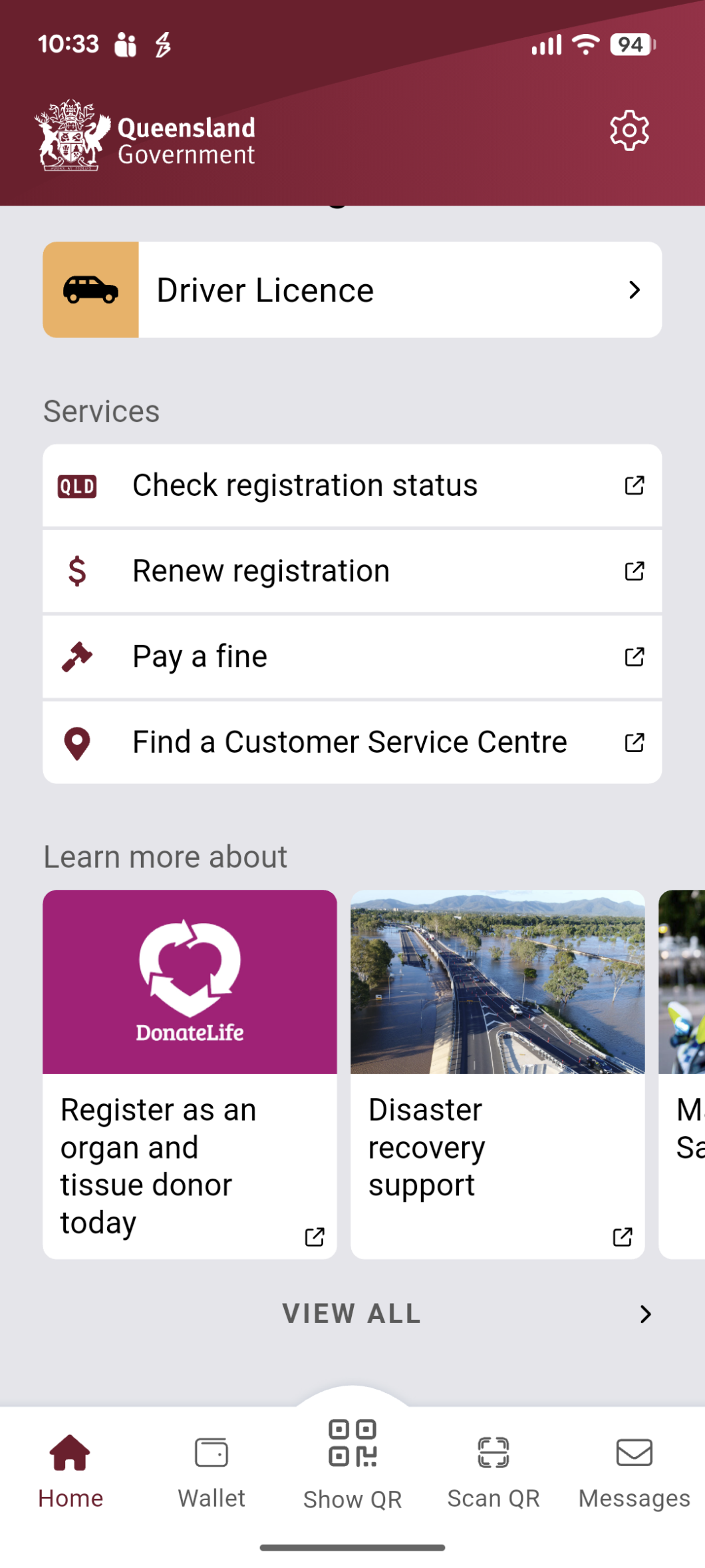Screenshot of a Queensland Government mobile app home screen showing a ‘Driver Licence’ card at the top and a list of services including ‘Check registration status,’ ‘Renew registration,’ ‘Pay a fine’, and ‘Find a Customer Service Centre’. Below, a ‘Learn more about’ section includes a DonateLife tile reading ‘Register as an organ and tissue donor today’, alongside a ‘Disaster recovery support’ tile. A navigation bar at the bottom shows icons for Home, Wallet, Show QR, Scan QR, and Messages.