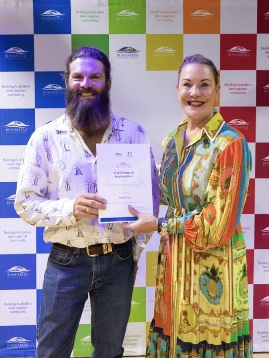 2 people stand in front of a colourful Bundaberg‑branded backdrop made up of blue, green, red, and orange panels. They are holding a framed Certificate of Nomination together. One person is wearing a patterned shirt and jeans, and the other is wearing a long, multi-coloured patterned dress.