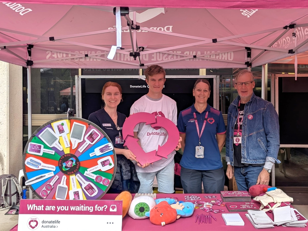 4 people stand under a pink DonateLife tent behind a table displaying promotional items like pens, brochures, and plush organs. A colourful prize wheel is visible on the left. The table features the text 'What are you waiting for?' and the Instagram handle '@donatelifeaustralia.' 