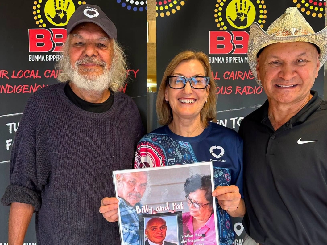 3 people stand in front of Bumma Bippera Media signage. One person holds a poster featuring a donor family story, while the group poses indoors during a community awareness activity supporting organ and tissue donation.