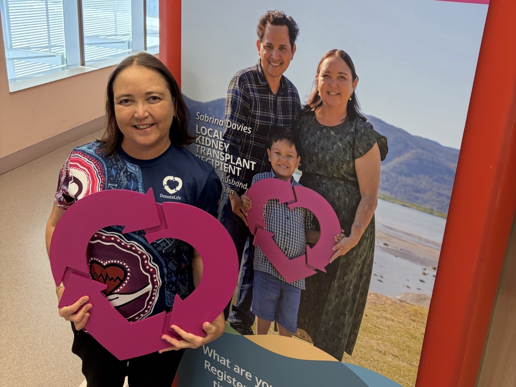 A person wearing a DonateLife shirt holds a large magenta DonateLife symbol while standing beside a promotional display featuring a family and information about organ donation and transplantation.