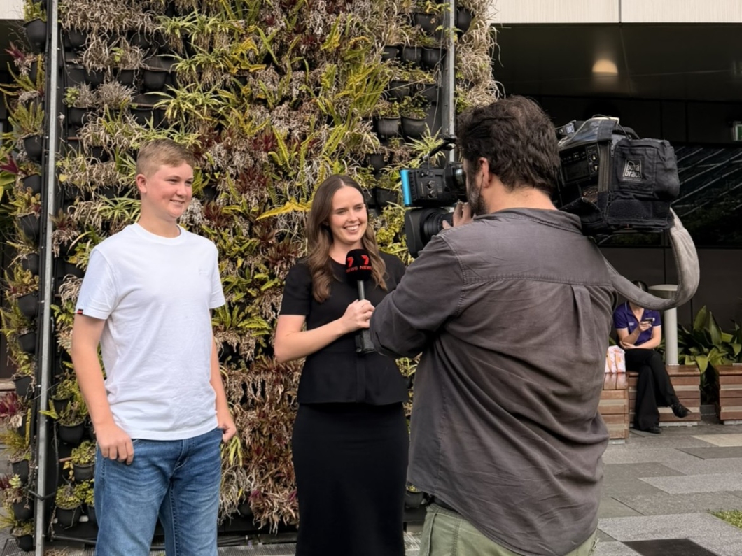 A television camera operator films an interview with 2 people standing in front of a large vertical garden wall. One person holds a Channel 7 microphone while speaking to camera, with the outdoor media interview taking place in a public plaza setting.