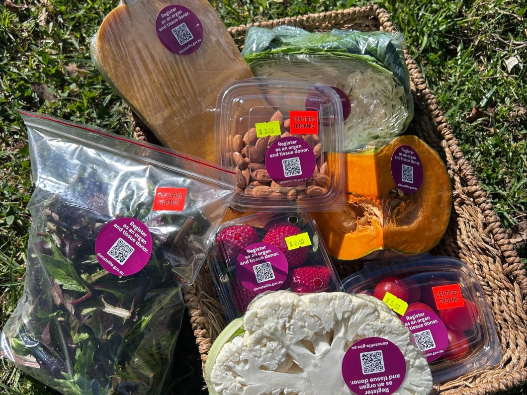 A woven basket filled with fresh organic produce sits on grass outdoors, including leafy greens, strawberries, almonds, tomatoes, cauliflower, pumpkin, cabbage and a wrapped loaf. Many of the items display circular magenta stickers featuring a QR code and the message ‘Register as an organ and tissue donor’. The produce is arranged in a colourful display promoting DonateLife registration through a local organic food business.