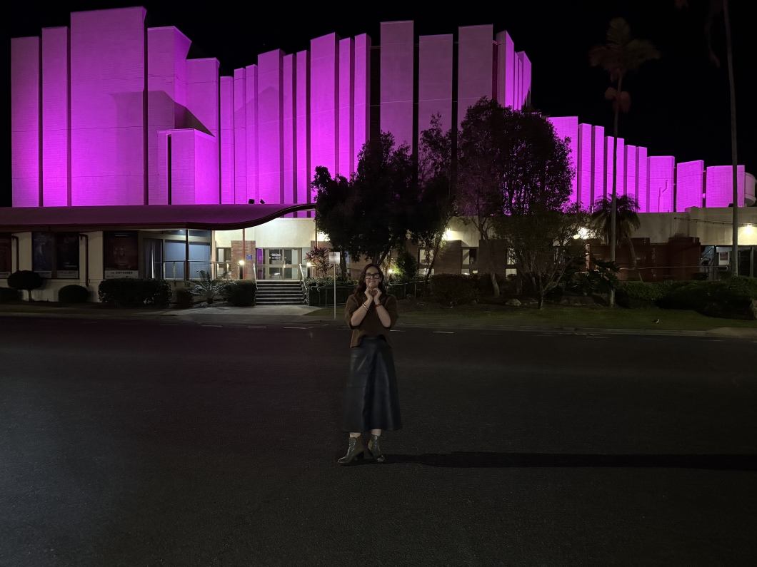 Pilbeam Theatre illuminated in magenta at night, with a person standing across the road in front of the building during a DonateLife awareness display.