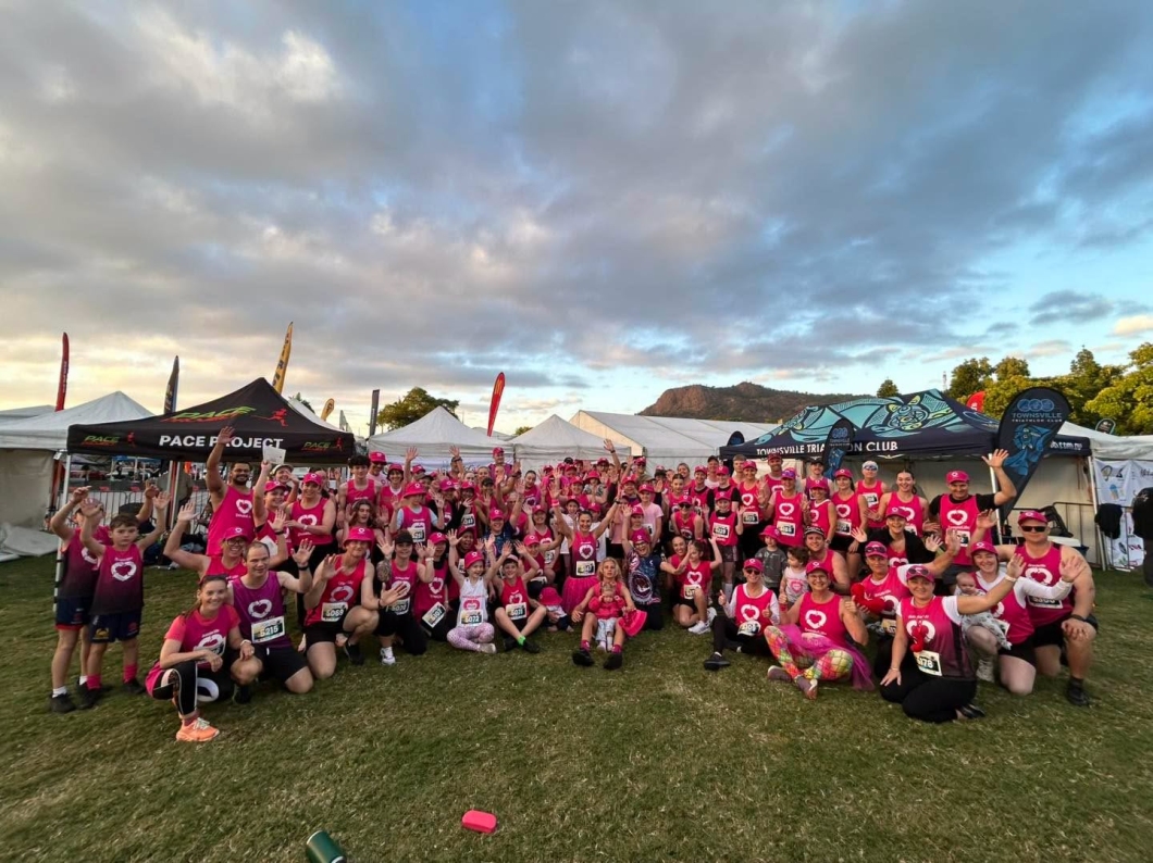 A large group of DonateLife supporters wearing magenta shirts, singlets and hats gather for a group photo at the Townsville Running Festival. Participants of all ages wave to the camera while standing on a grassed area surrounded by event marquees, with Castle Hill visible in the background.