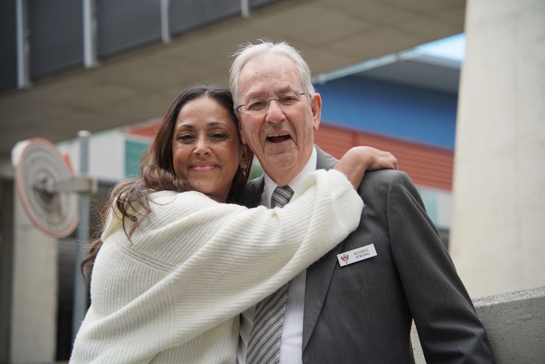 2 people standing close together outdoors, one wearing a white sweater and the other in a grey suit with a striped tie and a name tag reading 'RUSSELL STRONG'. The background features concrete structures and a building with blue and red panels.