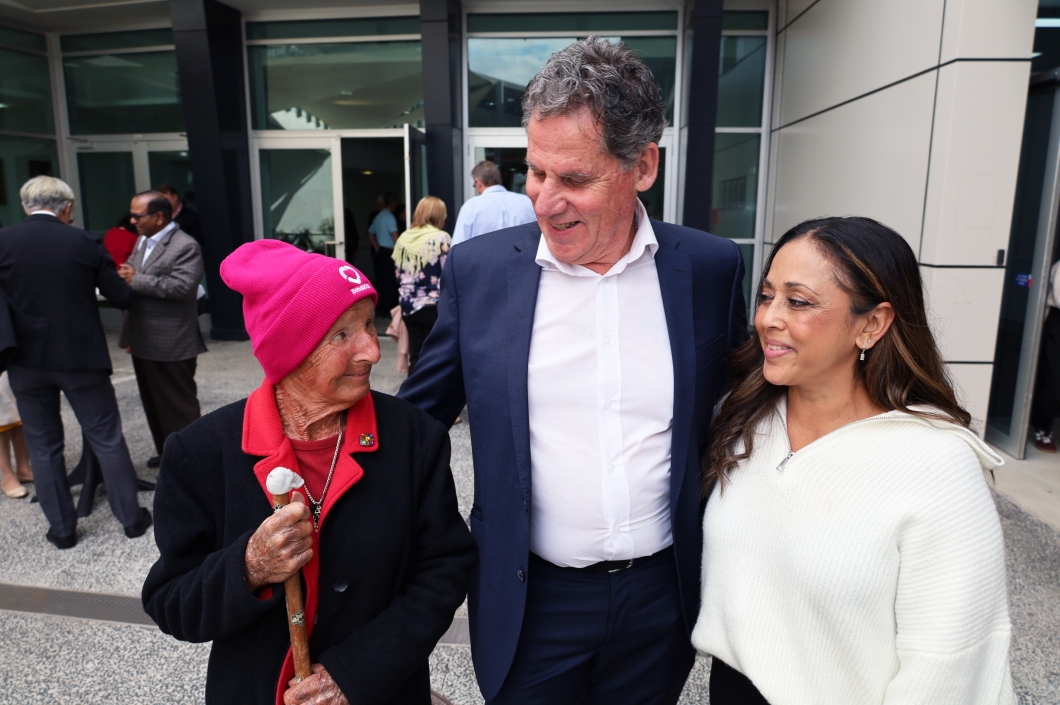3 people standing outside a building. The person on the left wears a pink beanie and holds a walking stick, the middle person wears a dark suit, and the person on the right wears a white sweater. Other people are visible in the background. 