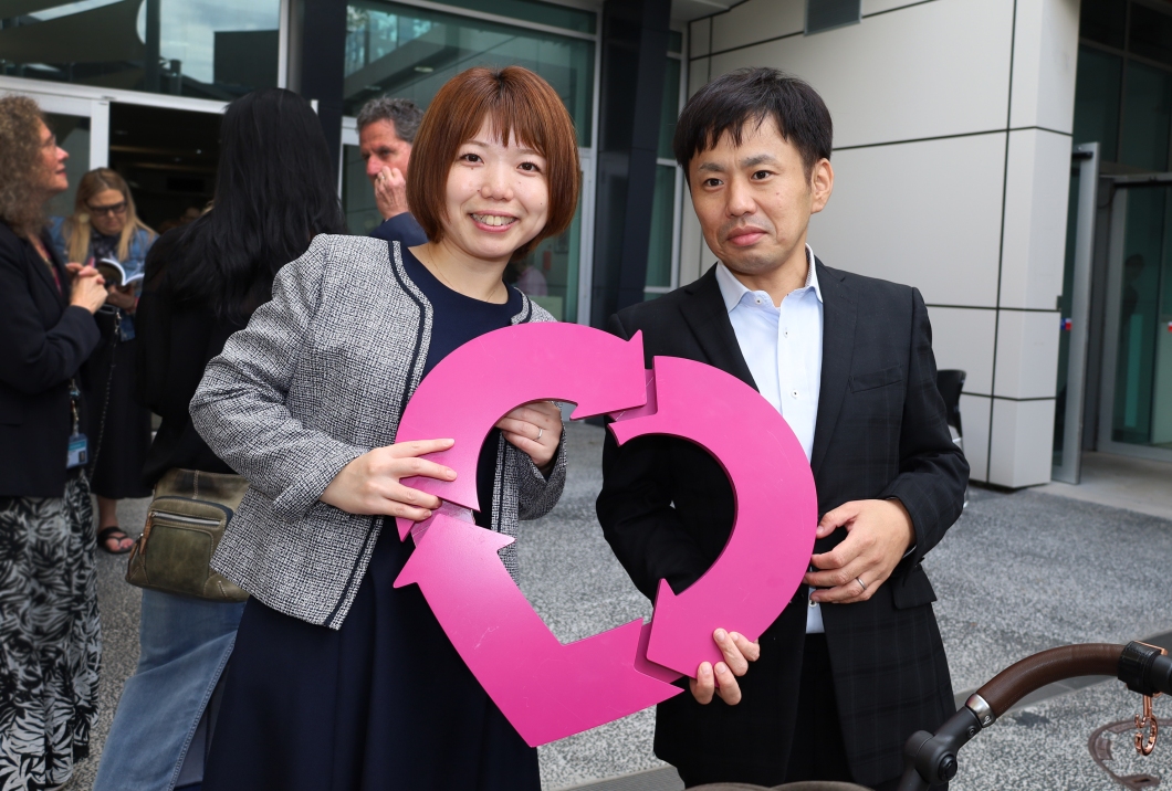 2 people standing outside a modern building holding a large DonateLife logo. Several other individuals are visible in the background, some engaged in conversation near the building entrance.