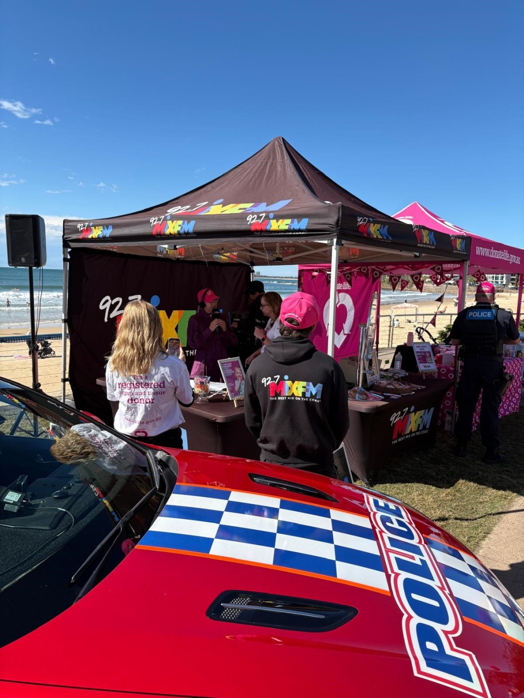 Promotional beach event featuring a tent with the ‘92.7 MIX FM’ logo and slogan ‘THE BEST MIX ON THE COAST’. People are gathered around, including someone in a branded hoodie and another in a shirt reading ‘registered organ and tissue donor’. A red police car with blue and white checkered markings is in the foreground, with ocean waves and sand in the background.