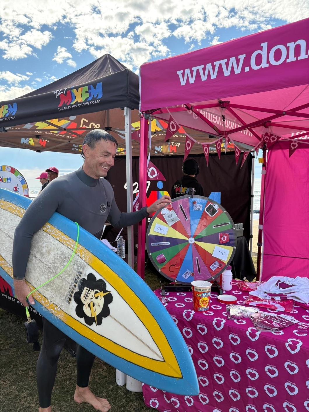 A person in a wetsuit holding a surfboard stands next to a colourful prize wheel at an outdoor event. The event is set up under black and pink tents with various promotional materials on the table, including flyers and shirts.