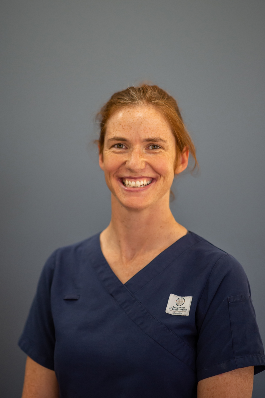 A person wearing navy clinical scrubs stands against a plain grey background. A small embroidered patch is visible on the left side of the scrub top. The person is shown from the chest up.