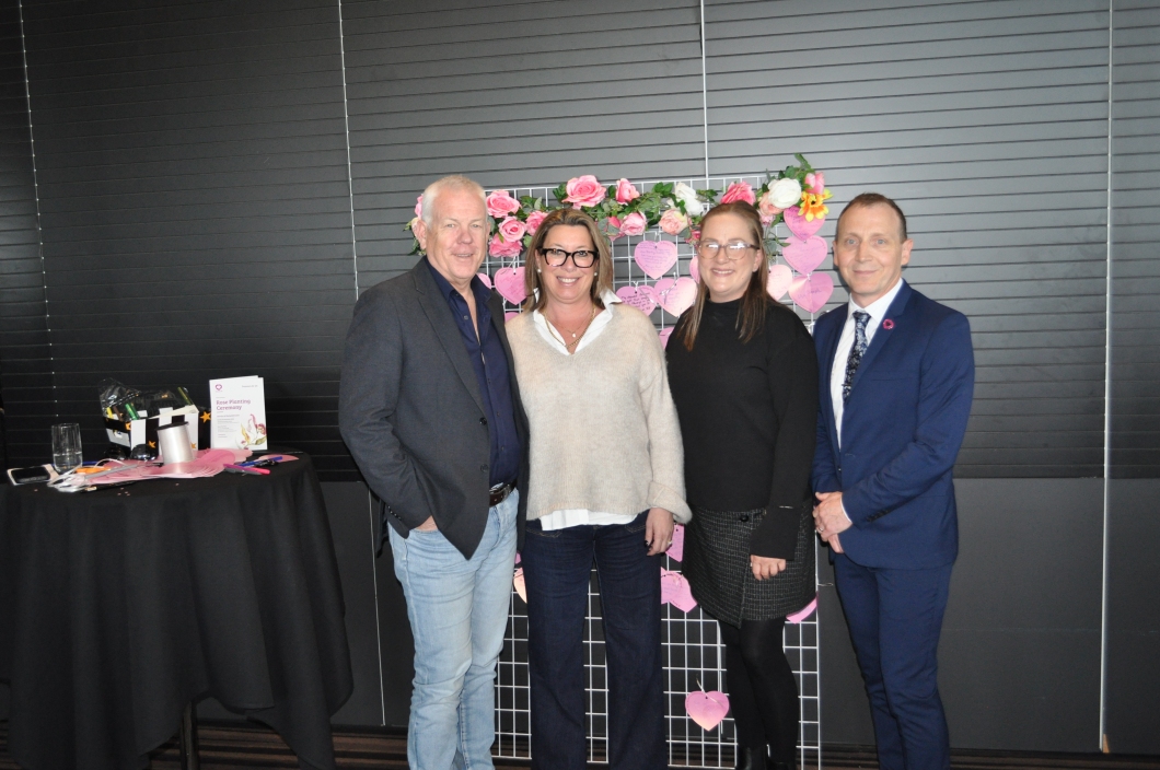 4 people stand together indoors in front of a decorative backdrop made of a white grid covered with pink paper hearts and artificial flowers. To the side, a tall round table holds event items including a glass, a booklet, and scattered pink paper hearts. The group is dressed in smart‑casual and formal clothing, and the background features dark horizontal wall panels.