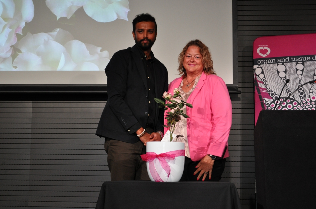 Two people stand behind a small table during a ceremony, with a white pot containing a rose plant placed in front of them. The pot is decorated with a pink ribbon. One person is dressed in dark clothing and the other in a bright pink jacket. A large screen behind them shows an image of pale flowers, and a DonateLife banner is visible to the side.