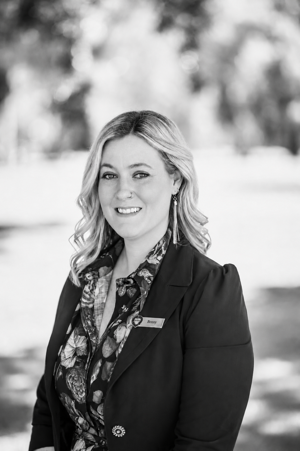 Black-and-white portrait of a DonateLife team member wearing a name badge and blazer, standing outdoors with trees softly blurred in the background.
