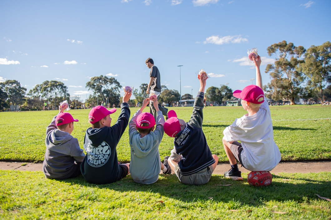 A group of children wearing magenta DonateLife caps sit on the edge of a sports field, raising their hands during the Charlie Stevens Memorial Match community event.