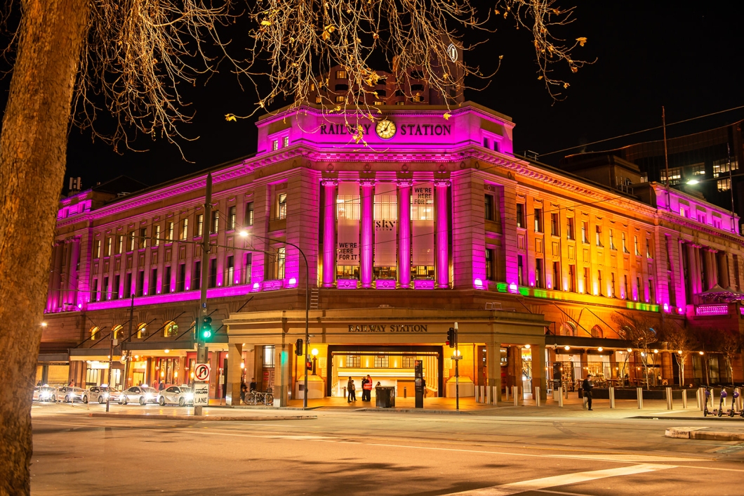 The historic Adelaide Railway Station is illuminated in bright magenta at night. The heritage building's façade, columns and clock tower glow against the dark sky, while streetlights, traffic signals and passing vehicles line the foreground. Bare tree branches frame the scene from above.