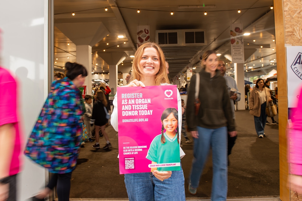 A community member stands at the entrance to a busy market holding a magenta DonateLife poster encouraging people to register as organ and tissue donors, while visitors walk past in the background.