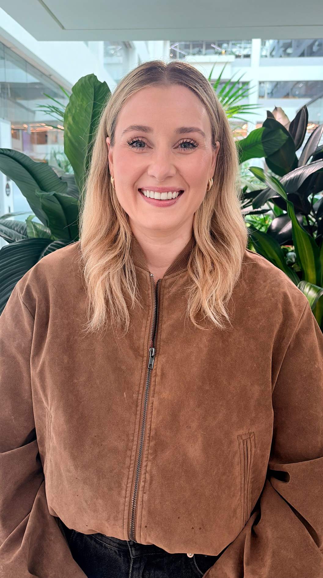 Portrait of a person wearing a brown jacket, standing indoors in front of large green plants in a bright office atrium.
