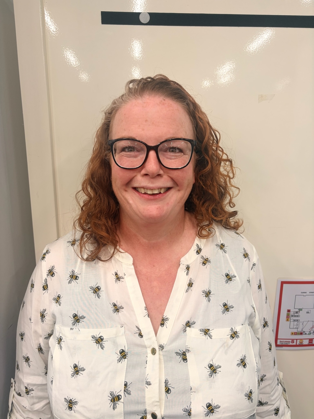 A person wearing glasses and a white shirt with a bee pattern poses for a portrait against a plain indoor background.