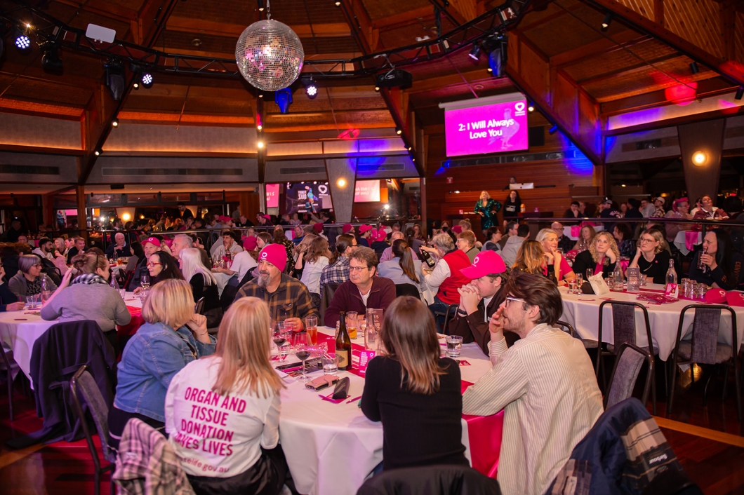 A large crowd gathers around round tables inside a function venue decorated with magenta DonateLife branding during a community quiz night and organ and tissue donation awareness event.