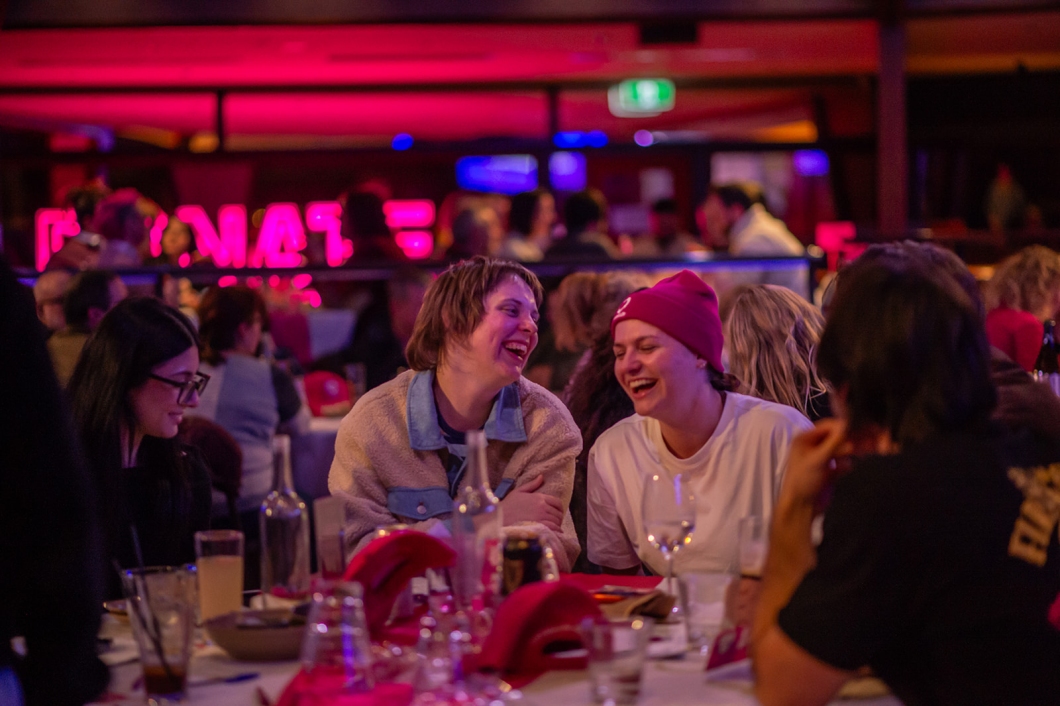 Guests sit together at a table during a DonateLife quiz night, with several attendees wearing magenta beanies and caps in a venue illuminated with magenta lighting.