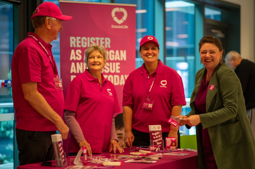 DonateLife volunteers and supporters stand at an information stall displaying registration materials and brochures, in front of a DonateLife banner encouraging people to register as organ and tissue donors.