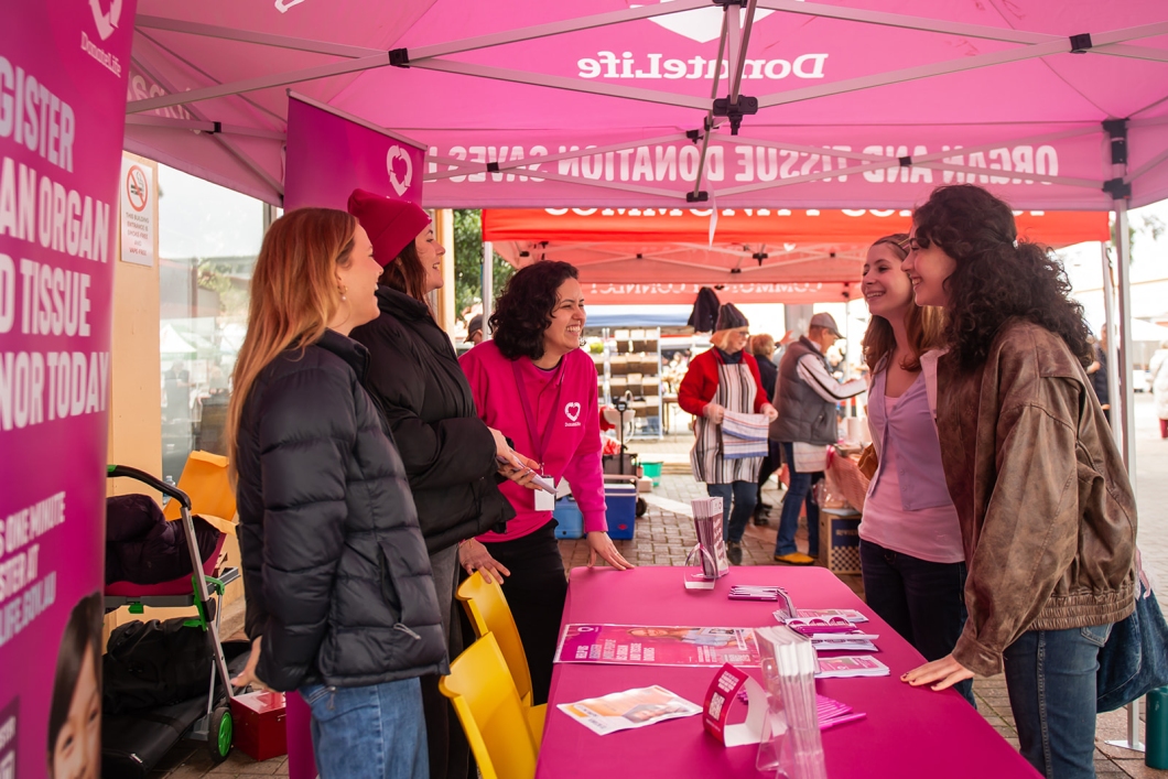 DonateLife representatives speak with visitors at an information stall under a magenta DonateLife marquee at a farmers market, sharing information about organ and tissue donation and donor registration.