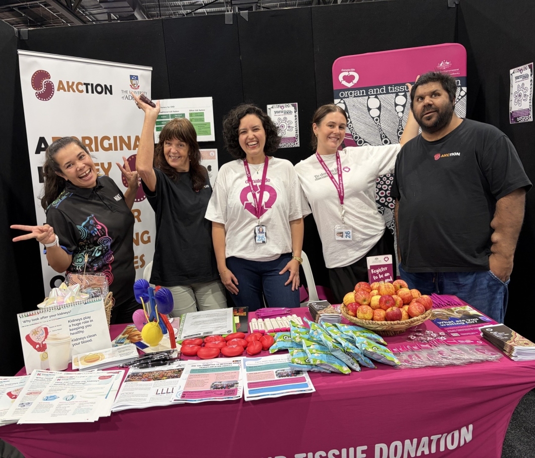 5 people stand behind a brightly coloured DonateLife information table at a community event. The table is covered with brochures, registration materials, pens, small giveaways, and a basket of apples, with a magenta DonateLife tablecloth and signage about organ and tissue donation visible. One person at the front gestures toward the camera while others stand side by side behind the display.